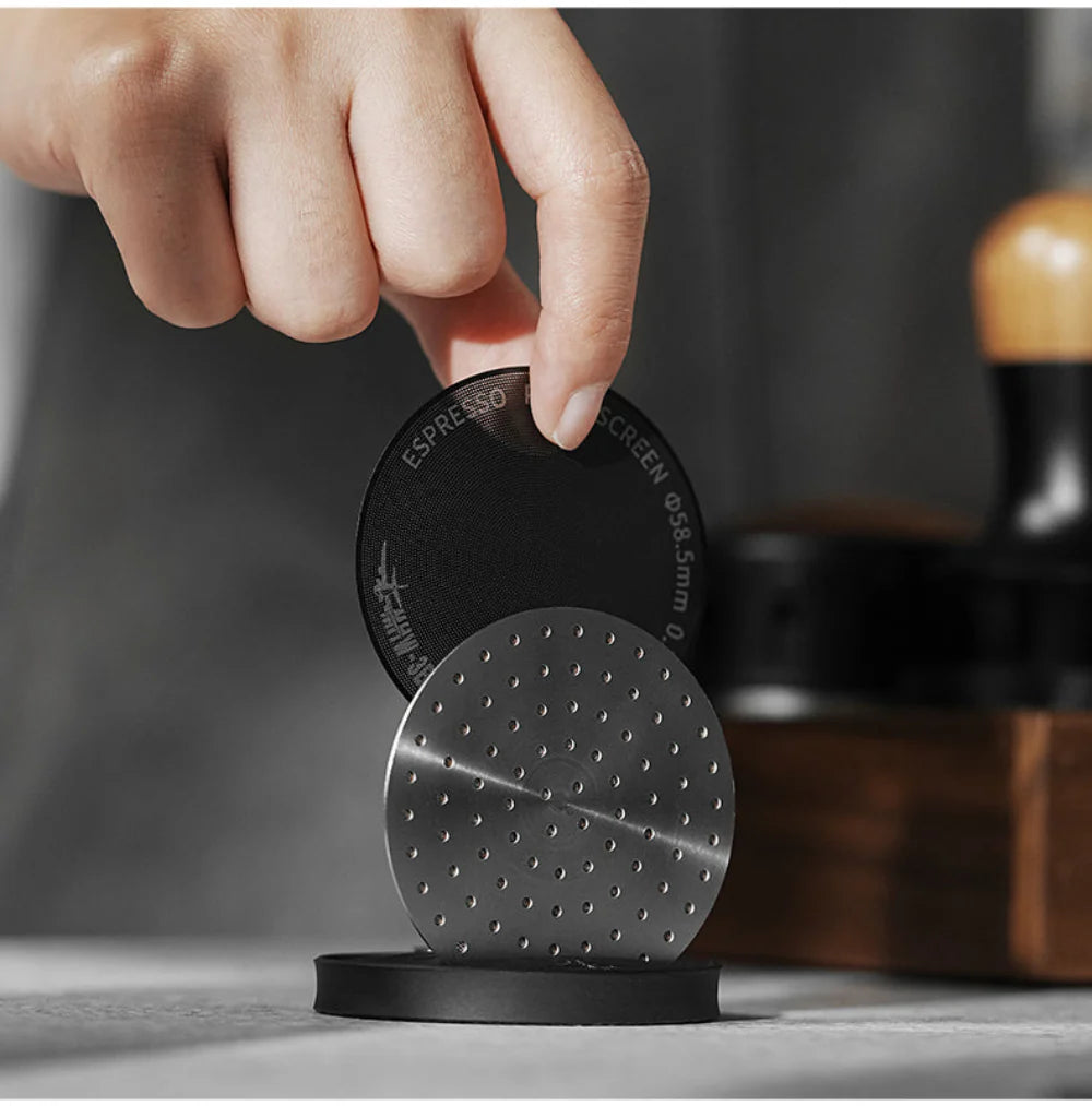 A hand holds the MHW-3BOMBER Espresso Puck Screen over a magnetic, sleek black holder with a wooden tamper blurred in the background on the gray countertop.