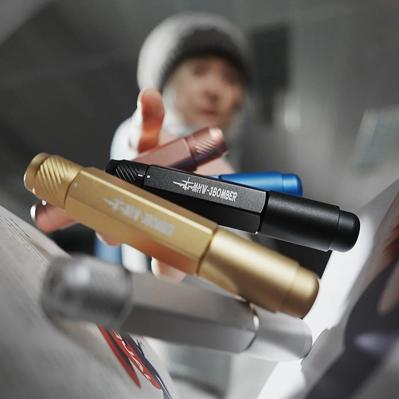 A hand reaches for vibrant MHW-3BOMBER Lightning Needle Distributor Pro WDT Tools with textured grips, suspended in mid-air and hinting at a telescopic design. In the blurred background, a person wearing a beanie is visible.