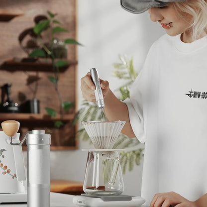 A person in a white shirt and cap uses a whisk in a glass pour-over coffee maker on a kitchen counter. Nearby is the MHW-3BOMBER Lightning Needle Distributor Pro WDT Tool and a thermos, with a plant in the background.