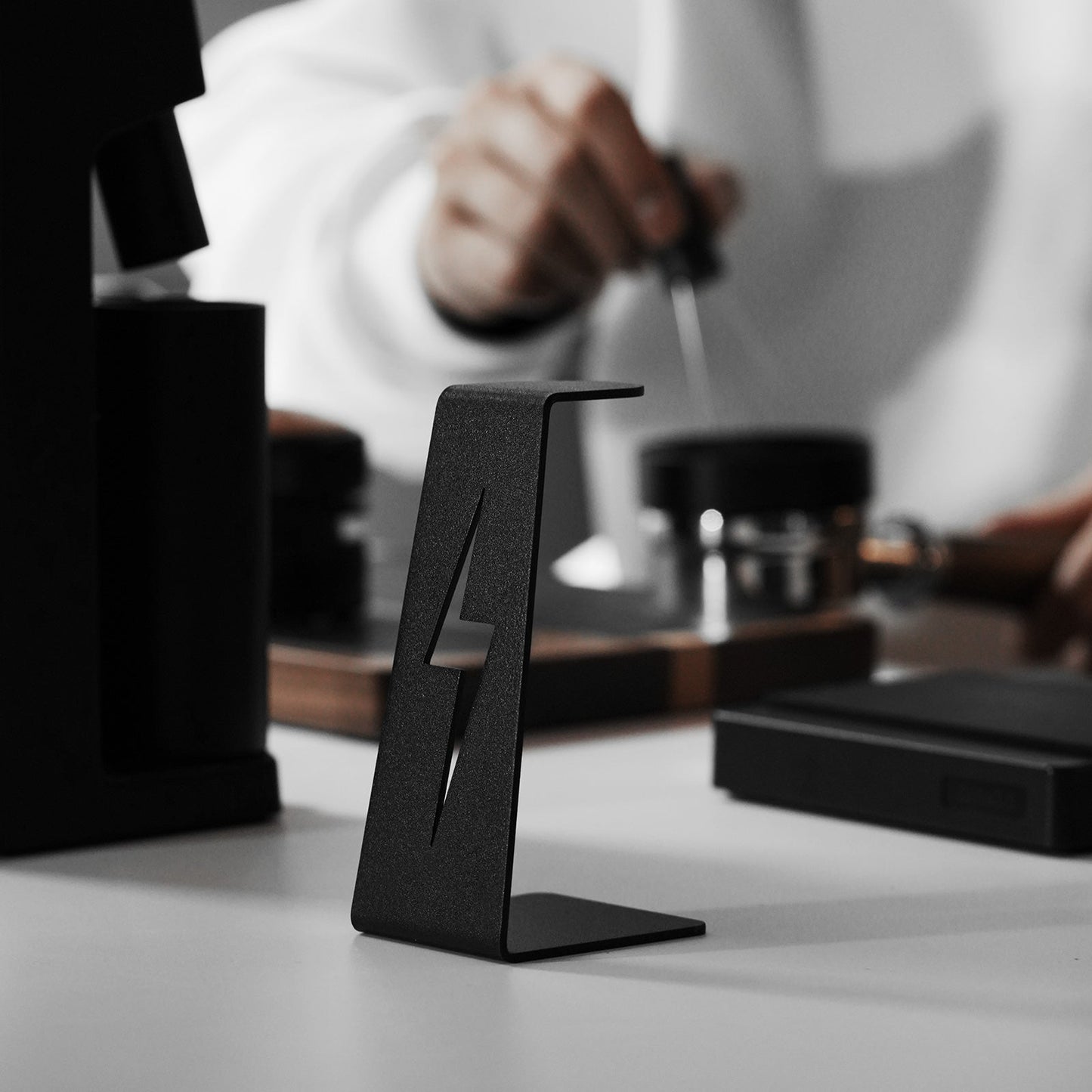 A close-up showcases the MHW-3BOMBER Lightning Needle Distributor WDT Tool 2.0, with its sleek black metal design, on a table. In the background, a person in a white shirt perfects coffee extraction using various tools, slightly blurred to add depth.