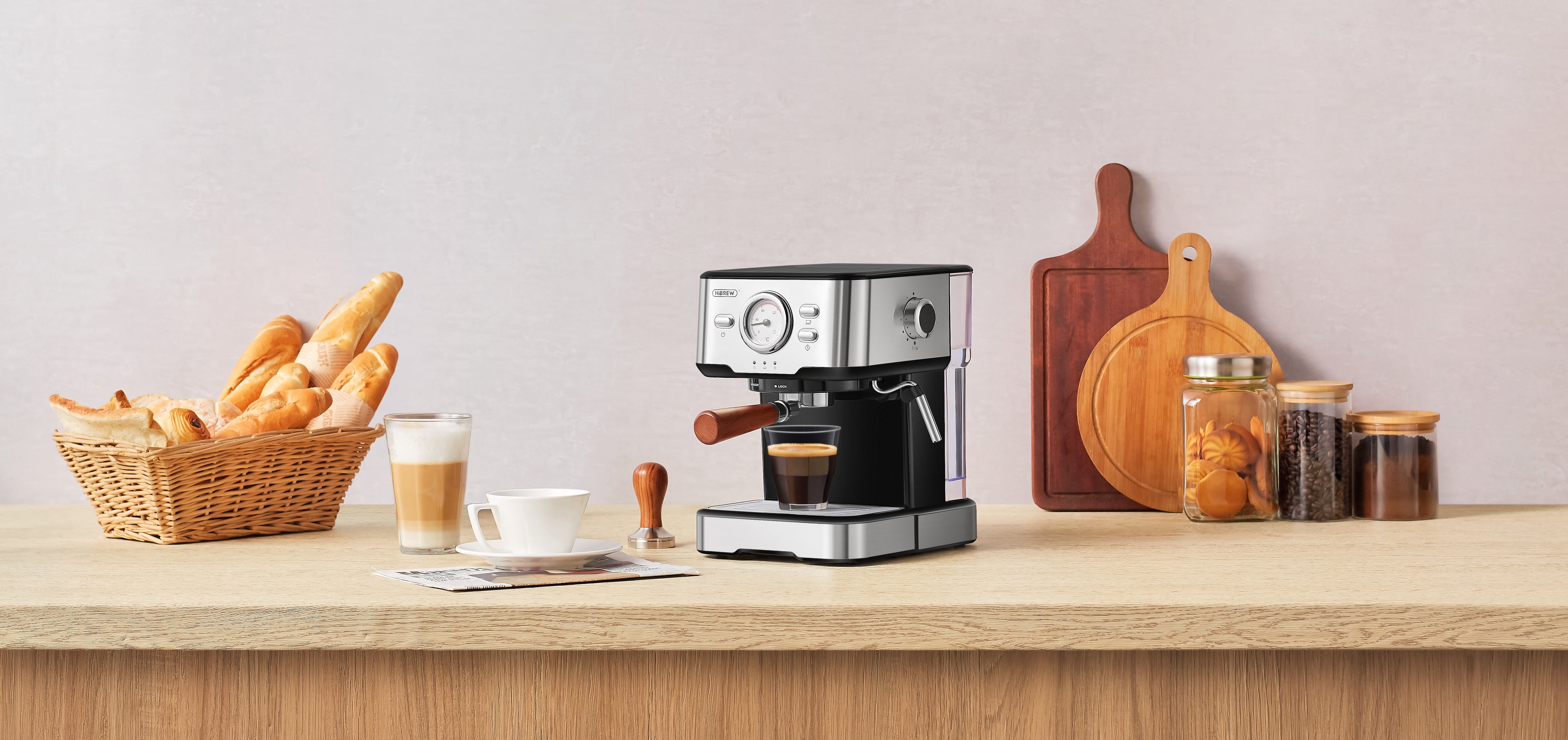 HiBREW espresso machine with wooden handle on a kitchen counter, alongside a cup of espresso, latte, tamper, and a basket of bread, showcasing a premium home coffee setup.