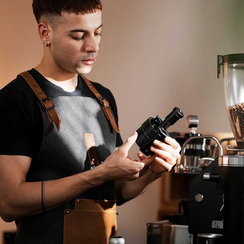 MHW-3BOMBER SE Pro WDT tool. Black distributor held by barista, near espresso machine.