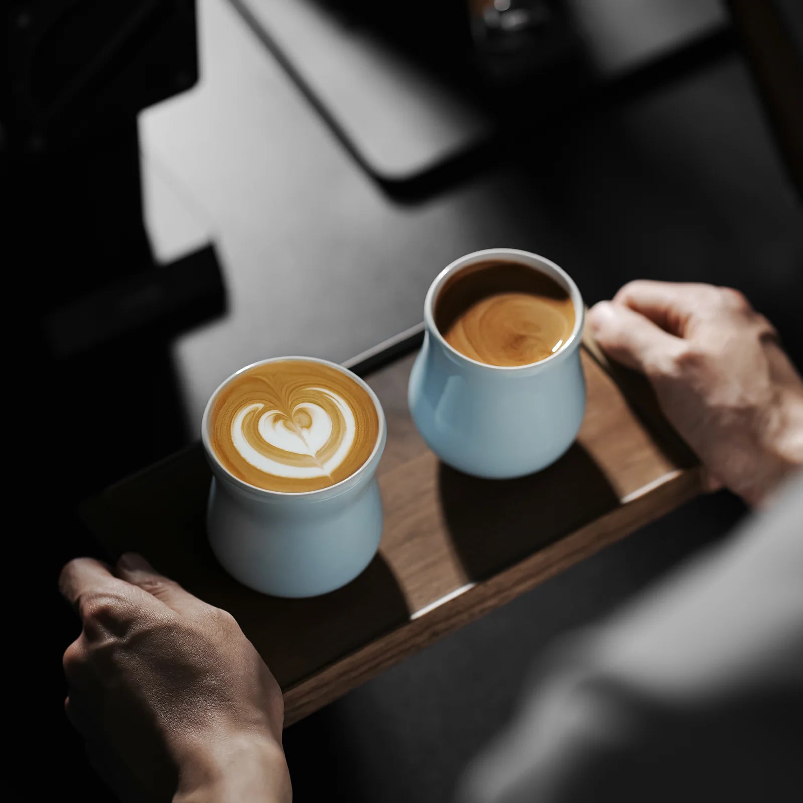 MHW-3BOMBER light blue ceramic cups with coffee, one showing latte art, on a wood tray.